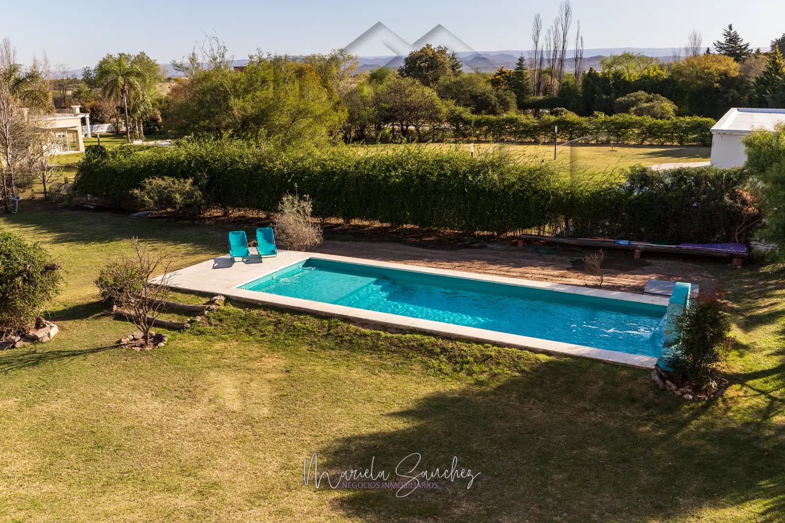 Vista aérea de la piscina rodeada de parque, con el horizonte serrano al fondo